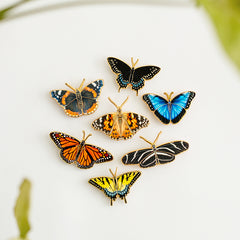 Seven butterfly enamel pins laid against a white background showing Blue Morpho, Black Swallowtail, Eastern Tiger Swallowtail, Monarch, Zebra Longwing, Painted Lady, and Red Admiral.