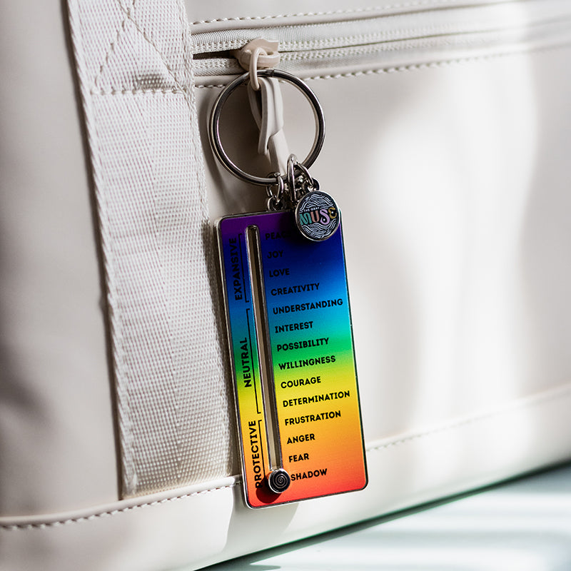 Rainbow gradient enamel keychain with a vertical frequency slider and a round music tag attached to a white backpack zipper.