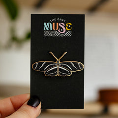 Zebra Longwing butterfly enamel pin on a black display card with The Gray Muse logo held by a hand