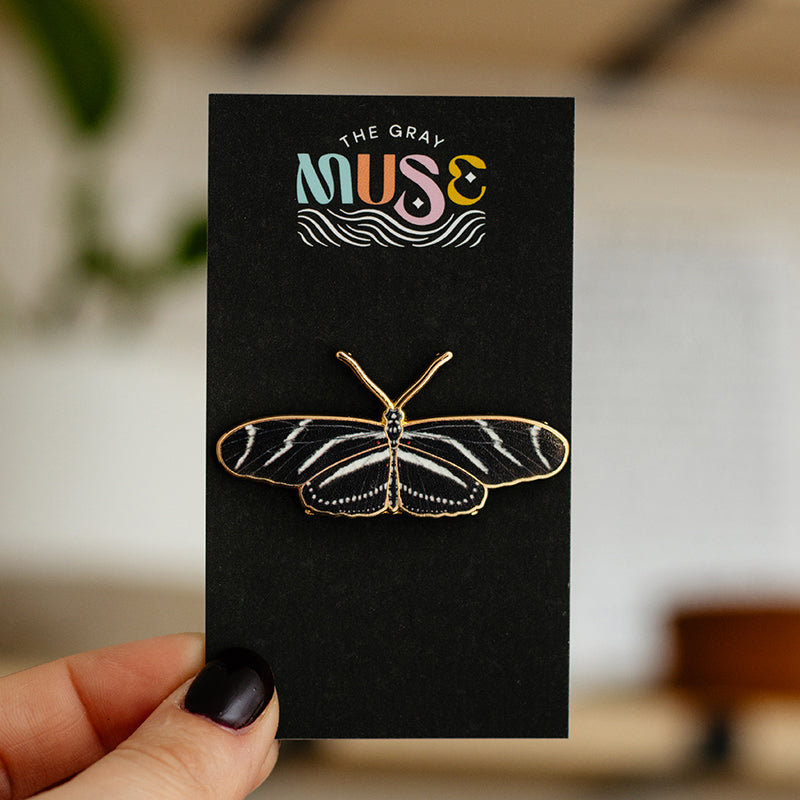 Zebra Longwing butterfly enamel pin on a black display card with The Gray Muse logo held by a hand