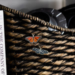 Two enamel butterfly pins on a woven basket, featuring an orange monarch and a dark blue pin with white vein markings