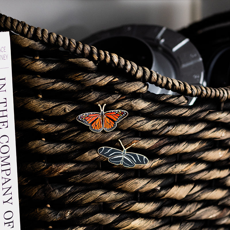 Two enamel butterfly pins on a woven basket, featuring an orange monarch and a dark blue pin with white vein markings