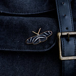 Zebra Longwing butterfly enamel pin on dark denim fabric beside a brass belt buckle