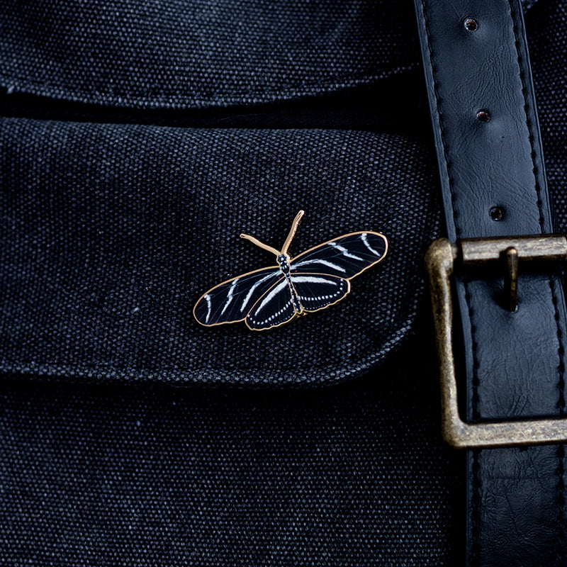 Zebra Longwing butterfly enamel pin on dark denim fabric beside a brass belt buckle
